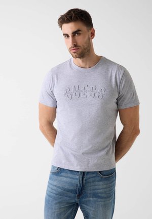 Mężczyzna o krótkich brązowych włosach, ubrany w szarą koszulkę "GUESS" i niebieskie dżinsy, stojący na białym tle.