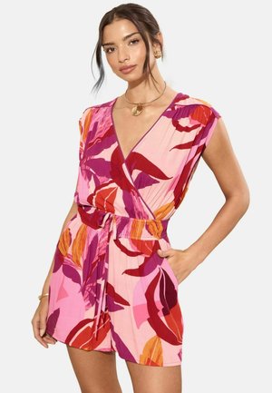 Femeie purtând o rochie tip petrecută cu modele florale în roz, roșu și portocaliu, cu mâneci scurte și cordon în talie, pozând cu o mână în buzunar și bijuterii aurii.