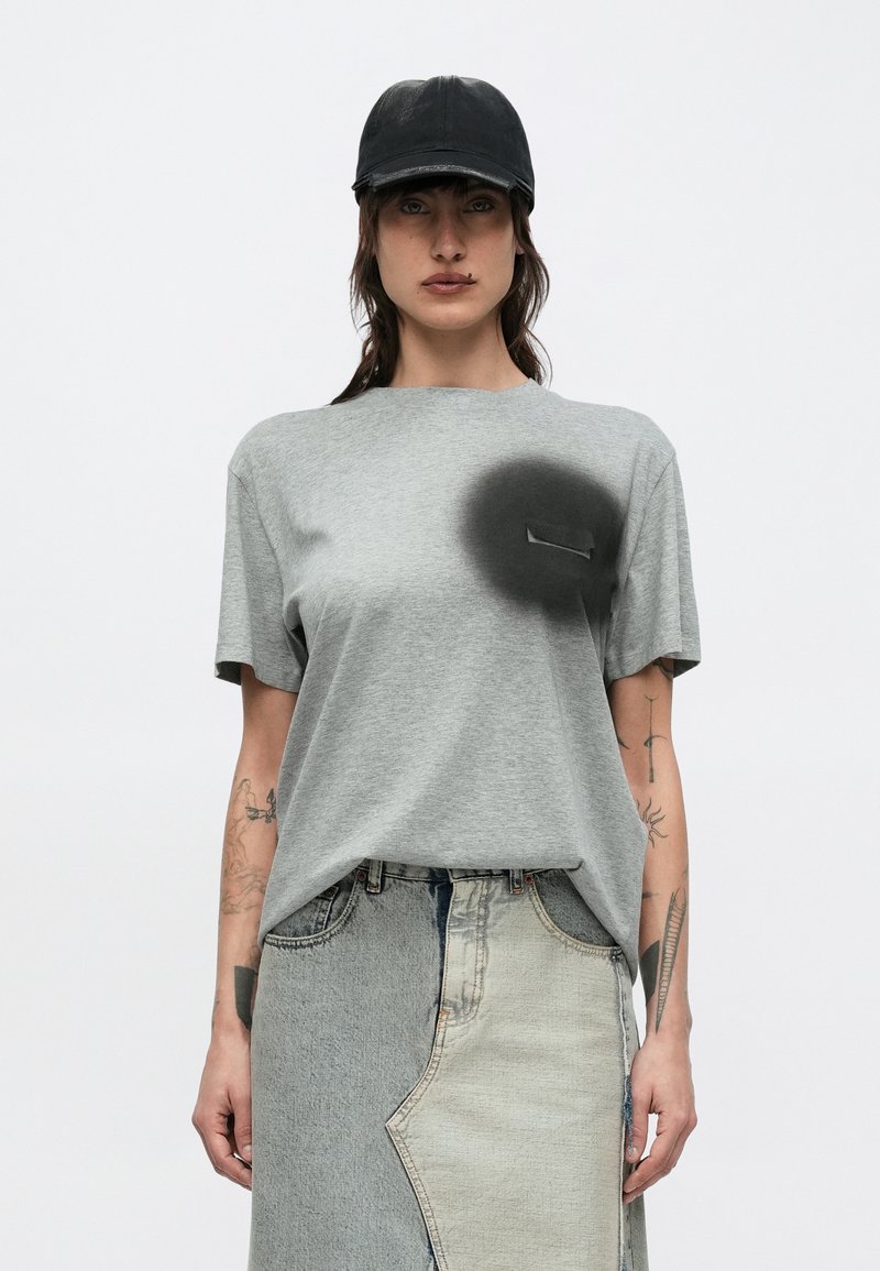 Naine, kellel on must nokamüts, hall t-särk musta spreivärvi detailiga ja kahetooniline teksaseelik, seisab ühevärvilise tausta eest.