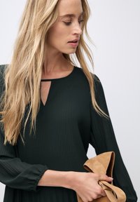 Dunkelgrünes geripptes Kleid mit einem Schlüssellochausschnitt und langen Ärmeln, gehalten mit einer tanfarbigen Wildleder-Clutch. Glatte Textur und fließendes Design.