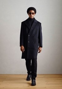 Zwarte dubbelzijdige jas met een brede revers, gedragen over een coltrui, gecombineerd met zwarte broeken en schoenen, op een model met zonnebril.