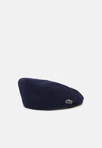Námořnicky modrý pletený baret se zaobleným tvarem a texturovaným povrchem, na boku je vyšívané zelené logo krokodýla.
