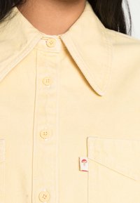 Gros plan sur une chemise boutonnée jaune pâle avec un col pointu et une petite étiquette en forme de champignon rouge sur la poche.