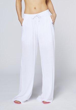 Woman wearing loose white pants with drawstring and hands in pockets, standing barefoot on a light background.