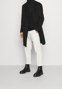 Abrigo negro de doble botonadura, suéter de punto oscuro con cuello alto, pantalones blancos y botas negras hasta el tobillo con detalles de costura. Texturas suaves y diseño ajustado.