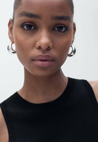 Close-up of a model's face with smooth skin, wearing silver hoop earrings and a black ribbed tank top. Neutral background highlights facial features.