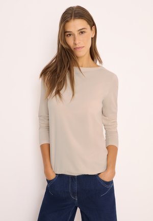 Haut beige à manches longues avec un col rond, texture lisse et coupe décontractée, assorti à un jean en denim bleu foncé avec des coutures visibles.