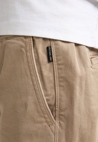 Pantalon beige en tissu texturé, avec des détails de couture et une étiquette noire portant l'inscription "PETROL IND" sur la poche avant.