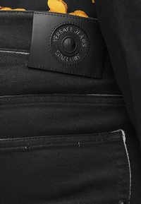 Melni denim džinsi ar teksturētu ādas birku, uz kuras ir iepresētas logo detaļas, novietota jostas vietā, ar redzamām izšuvuma detaļām gar kabatām.