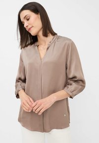 Silk-like taupe blouse with a v-neck, 3/4 puff sleeves, and a curved hem, featuring a subtle logo detail at the bottom.