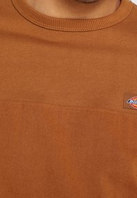 Hnědá bavlněná mikina s texturovaným povrchem, s žebrovaným kulatým výstřihem a malou nášivkou loga na levé straně.