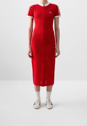 Personne portant une robe midi en maille rouge avec bordure blanche et fermeture éclair devant, associée à des chaussettes blanches et des baskets blanches à rayures noires.