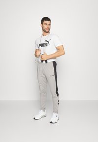 Vit kortärmad t-shirt med svart "PUMA"-logotyp, matchad med ljusgrå joggers med svarta sidpaneler och detaljer. Vita sportskor.