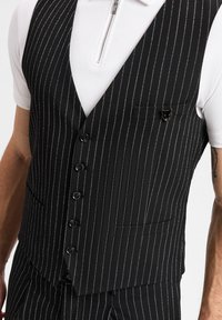 Schwarze Nadelstreifenweste mit silbernen Streifen, ausgestattet mit einer einzelnen Brusttasche, fünf vorderen Knöpfen und einem auffälligen bärenförmigen Accessoire.
