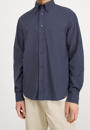 Mężczyzna ubrany w granatową koszulę zapinaną na guziki z długim rękawem oraz beżowe spodnie, stojący na tle jasnego, jednolitego tła.