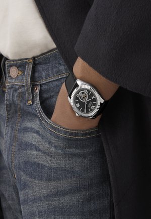 Silver stainless steel watch with a black leather strap, featuring a black dial, Roman numerals, and a visible movement. Worn on a wrist.