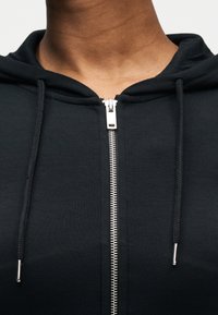 Gros plan sur une personne portant un sweat à capuche noir avec une fermeture éclair argentée et des cordons noirs avec embouts métalliques.