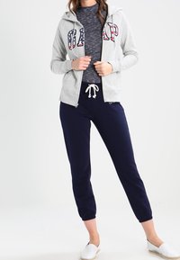 Sweat à capuche gris zippé avec le logo "GAP", pantalon bleu marine ajusté, et espadrilles blanches. Le sweat à capuche possède un cordon de serrage et une texture douce.