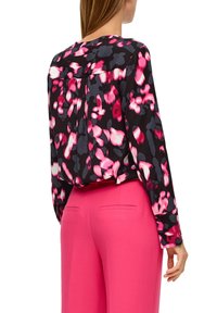 Mujer vestida con una blusa negra con un patrón floral abstracto en rosa y gris, metida dentro de pantalones sastre rosa brillante, vista desde atrás.