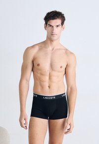 Zwarte boxershorts met een wit Lacoste-logo op de tailleband. De stof lijkt glad en rekbaar, waardoor ze nauwsluitend om het lichaam zitten.