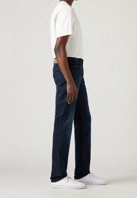 Homme portant un t-shirt blanc, un jean bleu foncé et des baskets blanches, debout de profil devant un fond uni.