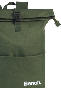 Sac à dos vert à rabat, fabriqué en tissu durable, avec une boucle noire, une surface texturée, une grande poche avant et un logo blanc.