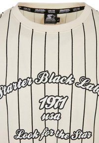 Cremefarvet T-shirt med sorte lodrette striber, der pryder teksten "Starter Black Label" og "1971 USA" i forskellige skrifttyper. Blød stofstruktur.