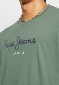 Oliwkowa bawełniana koszulka z okrągłym dekoltem, z luźnym krojem, z ciemnoniebieskim logo "Pepe Jeans" i napisem "LONDON" poniżej.