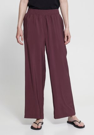 Persoană purtând pantaloni largi, largi, cu talie înaltă, de culoare burgund închis și papuci negri pe un fundal deschis.
