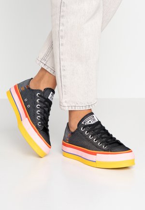 Person iført lyse jeans og sorte Converse-sneakers med flerfarvede tykke såler, stående på en hvid gulv.