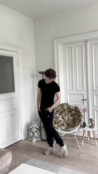 Black fitted t-shirt and black sweatpants; curly hair; light sneakers. Circular wicker chair with fur throw, patterned lanterns in background.