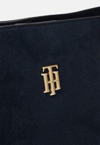 Logo dorato Tommy Hilfiger su tessuto scamosciato blu scuro, dettaglio ravvicinato di una scarpa o accessorio.