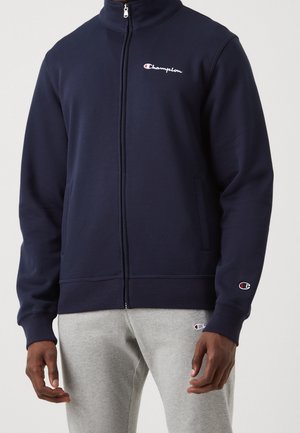 Mężczyzna ubrany w granatową bluzę Champion z suwakiem i jasnoszare spodnie dresowe z logo Champion na piersi, nadgarstku i udzie.