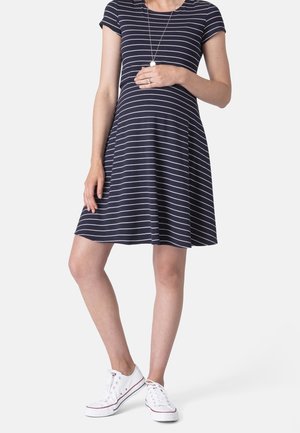 Zwangere vrouw in een marineblauwe en witgestreepte jurk, draagt witte sneakers en houdt haar buik met één hand vast.