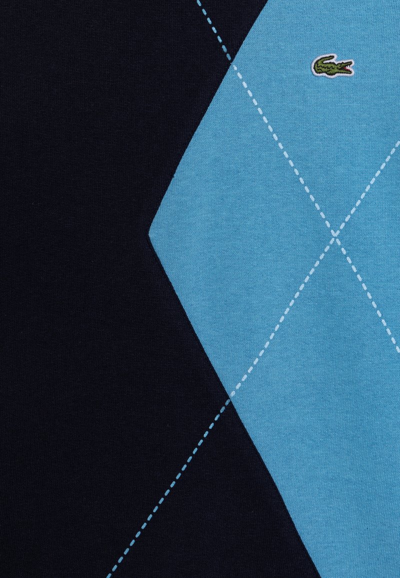 Pull en tricot avec un motif en losange bleu clair et marine, avec des coutures blanches et un logo de crocodile vert. Texture douce.