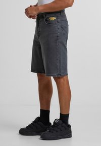 Shorts en denim gris avec une coupe droite, poches avant et arrière, et une patch de marque coloré. Associés à des baskets noires à lacets et des chaussettes.