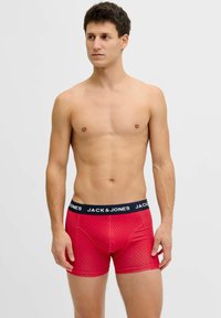 Vīrietis bez krekla ar īsiem tumšiem matiem, valkājot sarkanas rakstainas bokseršortus ar tumši zilu jostasvietu, uz kuras rakstīts "JACK & JONES", pret vienkāršu fonu.