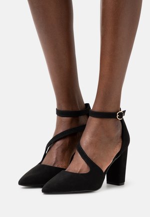 Chaussures à talons hauts en daim noir avec bouts pointus, brides croisées à la cheville et talon carré. Dotées d'une boucle dorée en accent sur la bride.