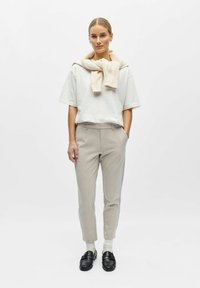 Femme portant un pantalon beige, une chemise à manches courtes blanche, un pull crème posé sur les épaules, des chaussettes blanches et des mocassins noirs, debout devant un fond blanc.