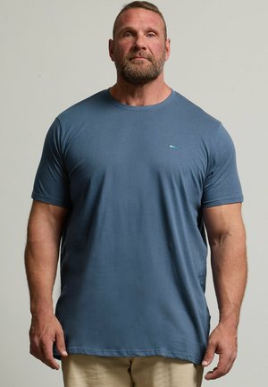 Uomo muscoloso con capelli castani corti e barba, che indossa una maglietta blu a maniche corte e pantaloni beige, in piedi davanti a uno sfondo semplice.