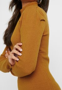 Žebrovaný hořčicově žlutý top s dlouhým rukávem, který má vysoký výstřih, přiléhavý siluetu a nabírané detaily na ramenech. Texturovaná tkanina.