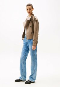 Manteau trench marron à double boutonnage avec épaulettes, chemise blanche, jeans larges bleu clair et mocassins noirs. Texture lisse, style décontracté.