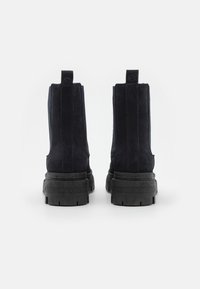 Botines de ante negros con suela gruesa, que cuentan con tiradores traseros y suelas de goma texturizada en contraste.