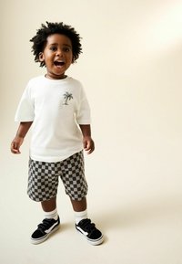 Weißes T-Shirt mit Palmenmotiv, karierten schwarzen und grauen Shorts und schwarzen Turnschuhen mit Klettverschlüssen, getragen mit weißen Socken.