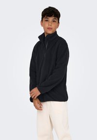 Svart fleece-pull med høy hals og kvart glidelås. Har lange ermer og en avslappet passform. Brukt over lyse bukser.