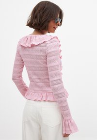 Roza čipkasta majica z ruffled ovratnikom in manšetami, dolgimi rokavi in peplum robom. Tkanina ima teksturiran, črtast vzorec.