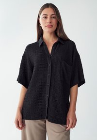 Camisa negra texturizada de manga corta, con botonadura frontal y un bolsillo en el pecho, fabricada con un tejido arrugado.