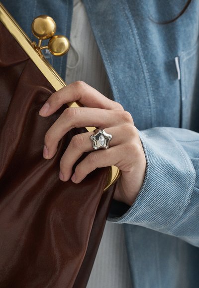 Roka ar sudraba zvaigznes formas gredzenu, tur brūnu ādas maku ar zelta aizdari, valkājot gaiši zilu džinsa jaku un baltu topu.