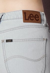 Jeans in denim azzurro con un'etichetta in pelle marrone impresso con "Lee" sulla vita, con cuciture sui taschini posteriori e un piccolo dettaglio dell'etichetta.