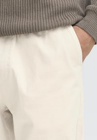 Light beige cotton trousers with a smooth texture, featuring side pockets and a subtle seam detailing at the sides. The waistband sits at the hips.